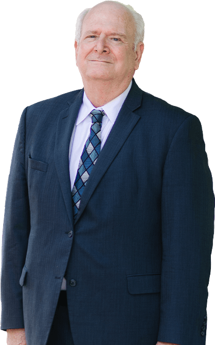 A man with dark hair and a beard, wearing a dark striped suit, light blue shirt, and pink tie, stands smiling with one hand in his pocket.