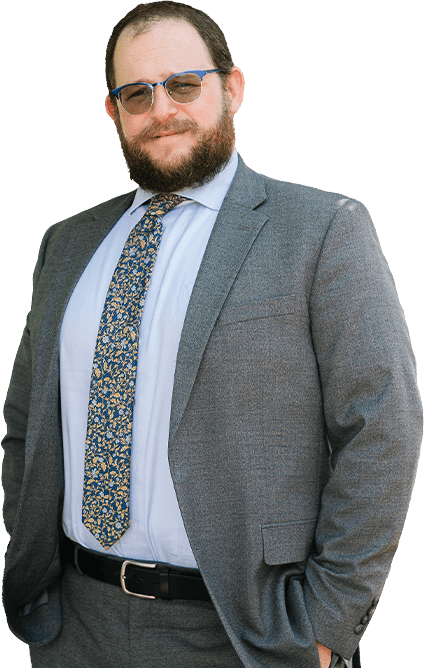 A man with dark hair and a beard, wearing a dark striped suit, light blue shirt, and pink tie, stands smiling with one hand in his pocket.
