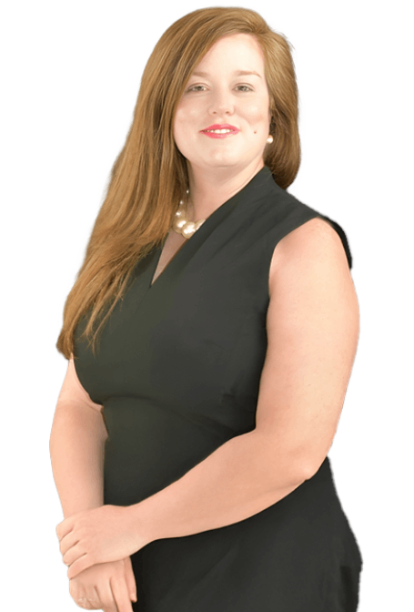 A woman with long brown hair wearing a sleeveless black dress and a pearl necklace stands smiling with her hands clasped in front of her against a white background.