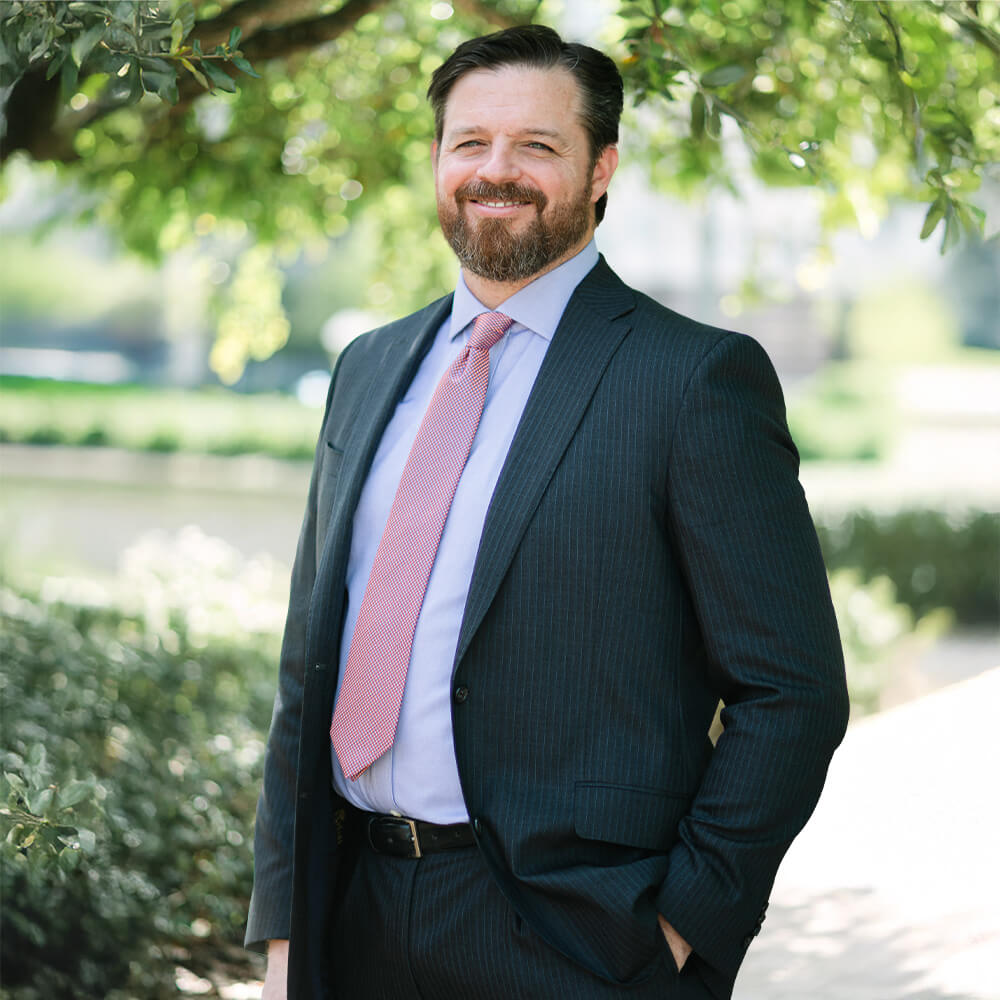 A man with a beard and mustache wearing a dark pinstripe suit, light blue shirt, and pink tie stands outdoors under a leafy tree, smiling with one hand in his pocket.