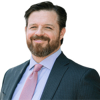 A man with short dark hair and a beard, wearing a dark suit, light blue shirt, and pink tie, is smiling in front of a white background.