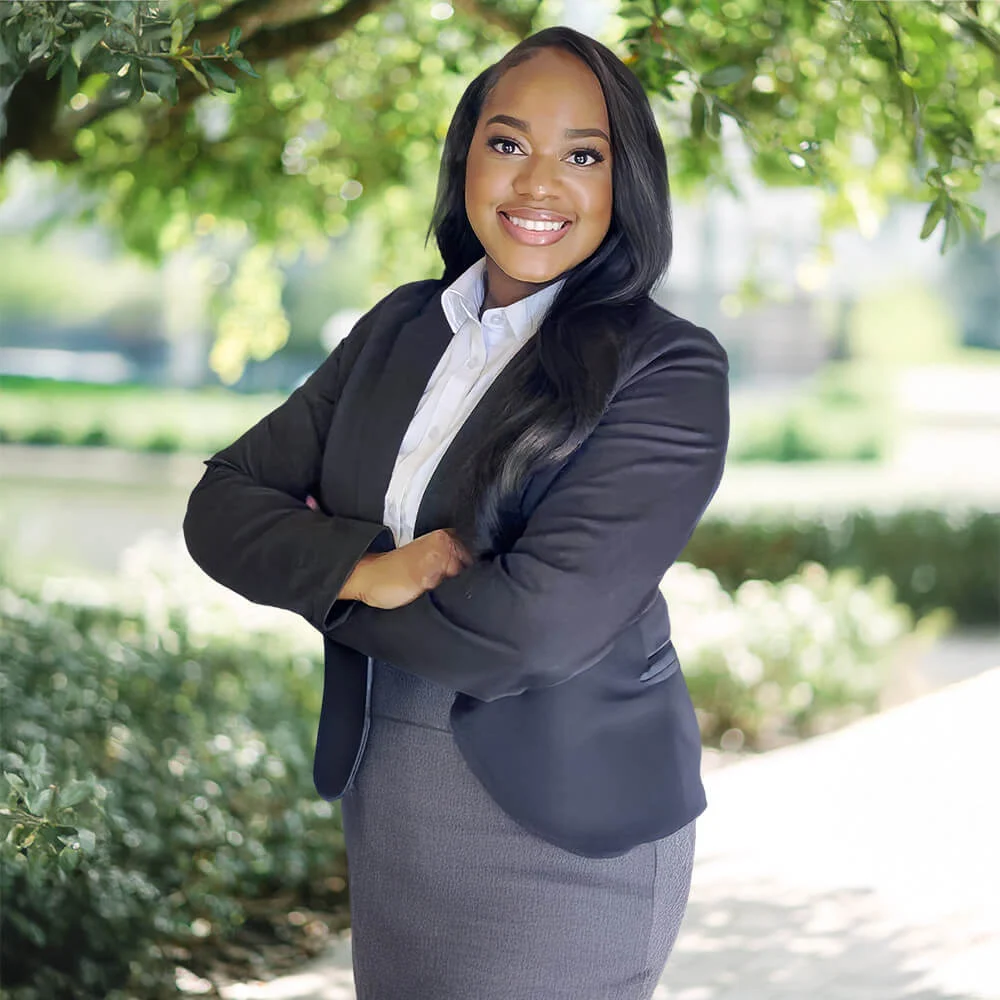A woman in professional attire stands outdoors under tree branches, smiling confidently with arms crossed. She wears a dark blazer and skirt, with a white blouse, and has long, straight hair. The background is bright and leafy.