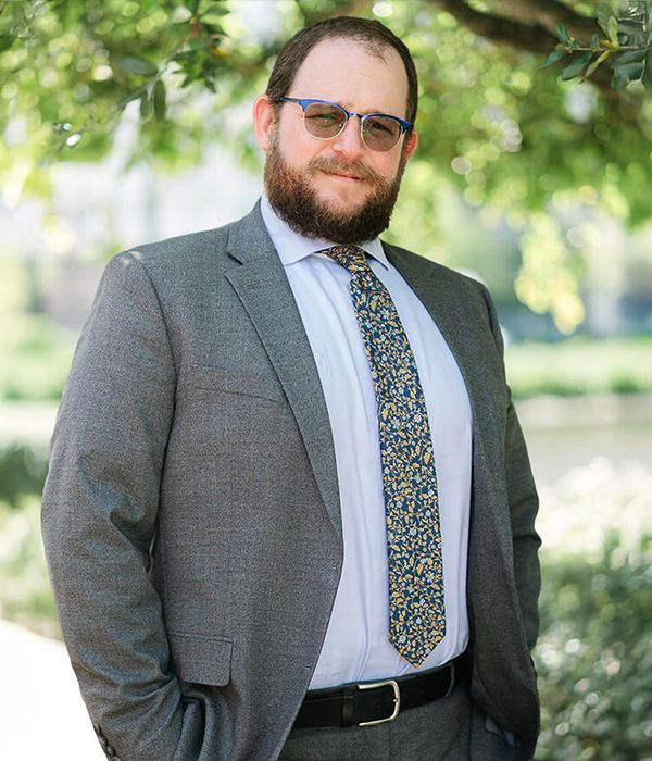 A man with a beard and sunglasses, wearing a gray suit, white shirt, and patterned tie, stands outdoors in front of leafy green trees, smiling slightly at the camera.