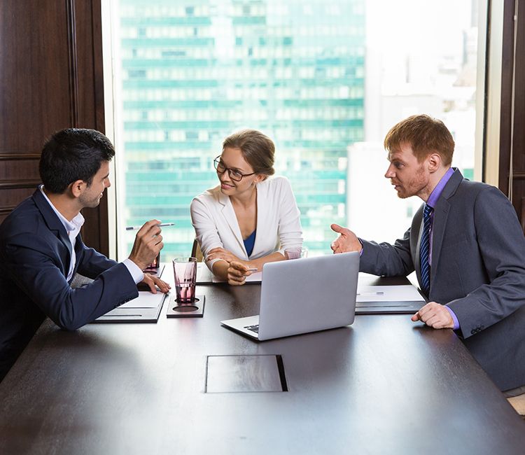 Three business professionals sit around a table in an office, engaged in a discussion. Two men and one woman are talking, with a laptop and documents in front of them. A large window shows a cityscape in the background.