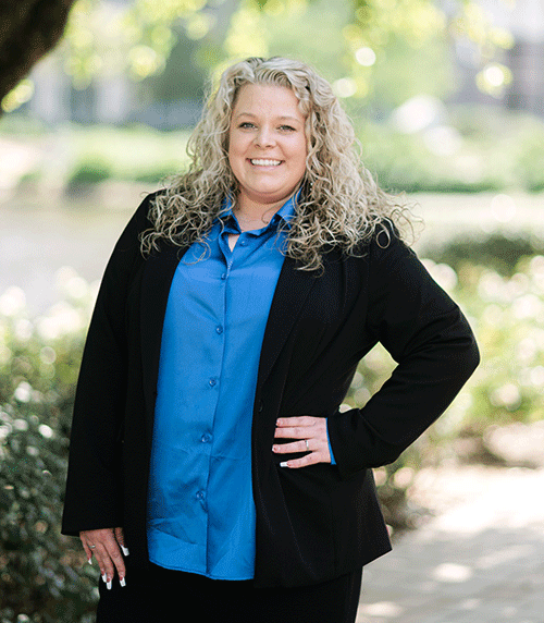 A woman with long, curly blonde hair stands outdoors, smiling. She wears a bright blue button-up shirt under a black blazer and has one hand on her hip. The background is leafy and sunlit, with greenery and blurred trees.
