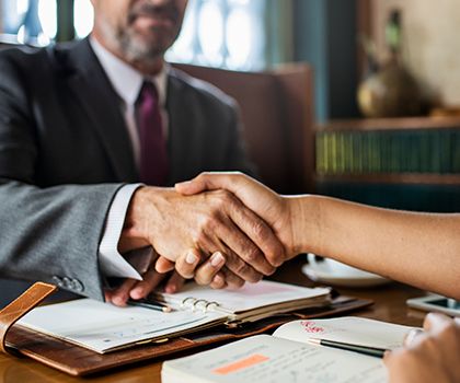 A person in a suit shakes hands with another person across a desk, with notebooks and documents open in front of them, suggesting a business meeting or agreement.