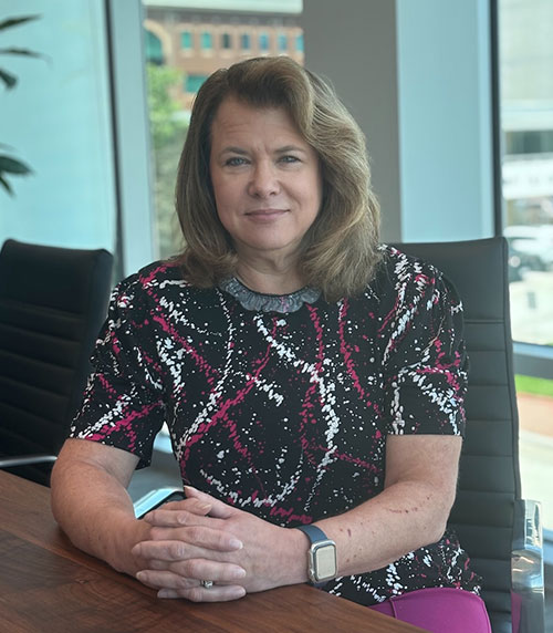 A woman with shoulder-length brown hair sits at a conference table, hands folded, wearing a black, white, and pink patterned top and a smartwatch, with large windows and office chairs in the background.