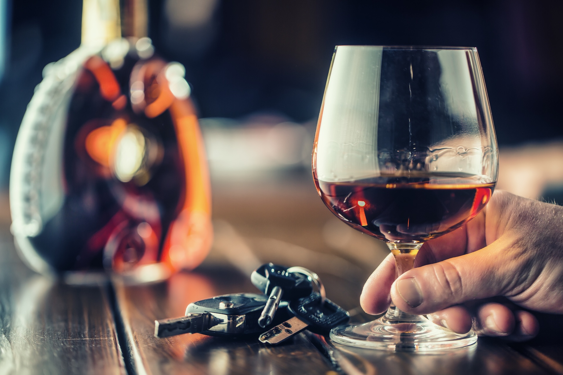 A hand holding a glass of amber alcohol sits next to car keys on a wooden table, with a blurred liquor bottle in the background, suggesting the risks of drinking and driving.