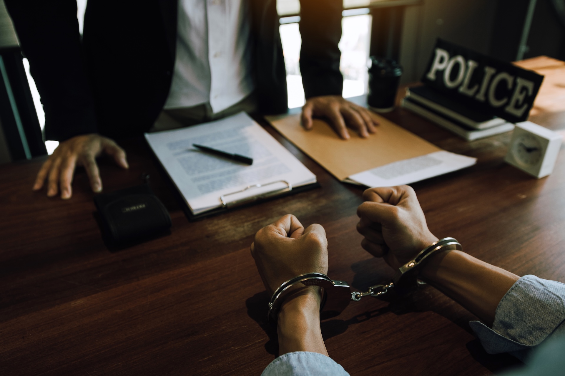A person in handcuffs sits across a desk from an officer in an office, with documents, a pen, and a "POLICE" sign visible on the wooden table.
