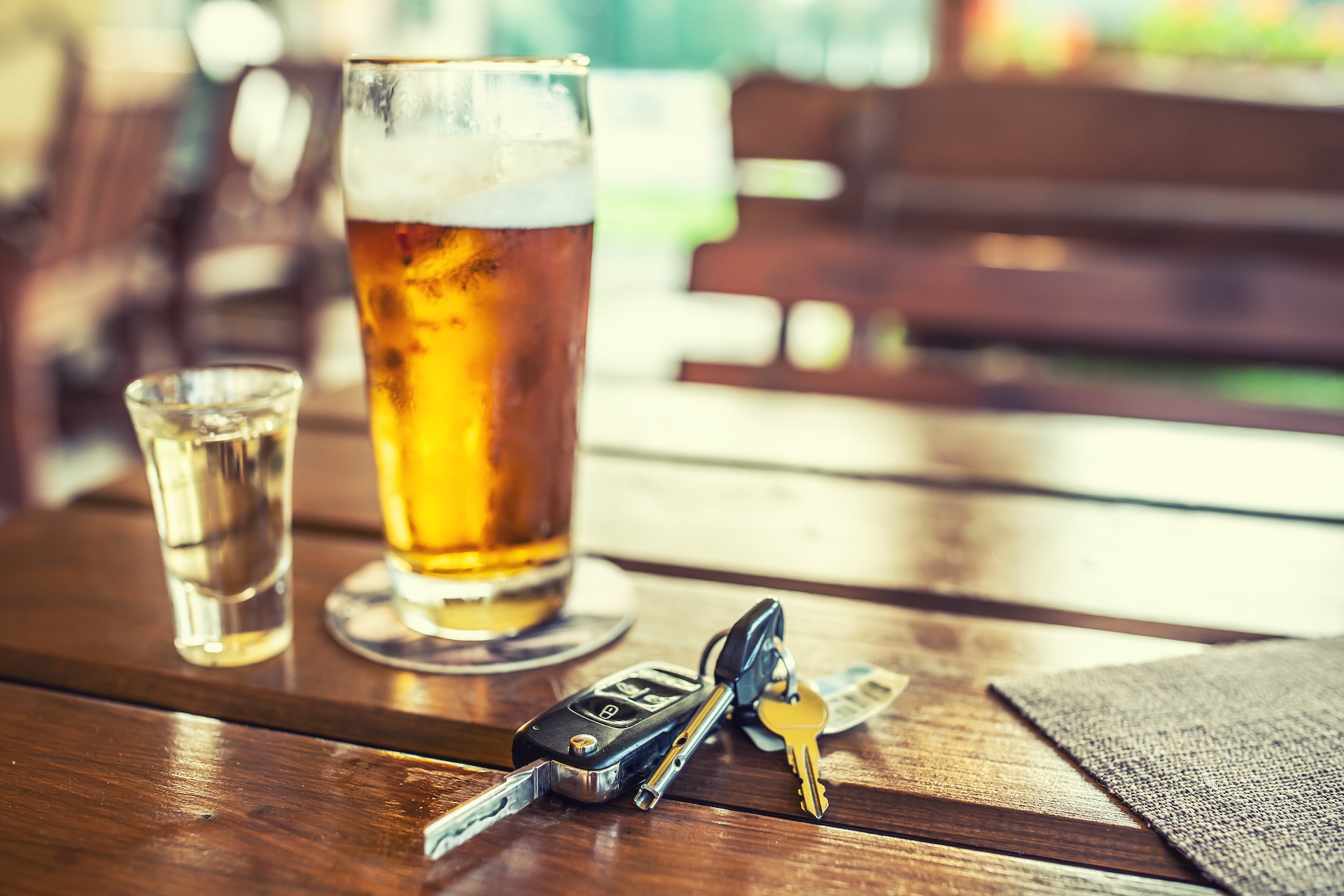 A glass of beer, a shot of liquor, and a set of car keys are on a wooden table in an outdoor seating area, suggesting the dangers of drinking and driving.