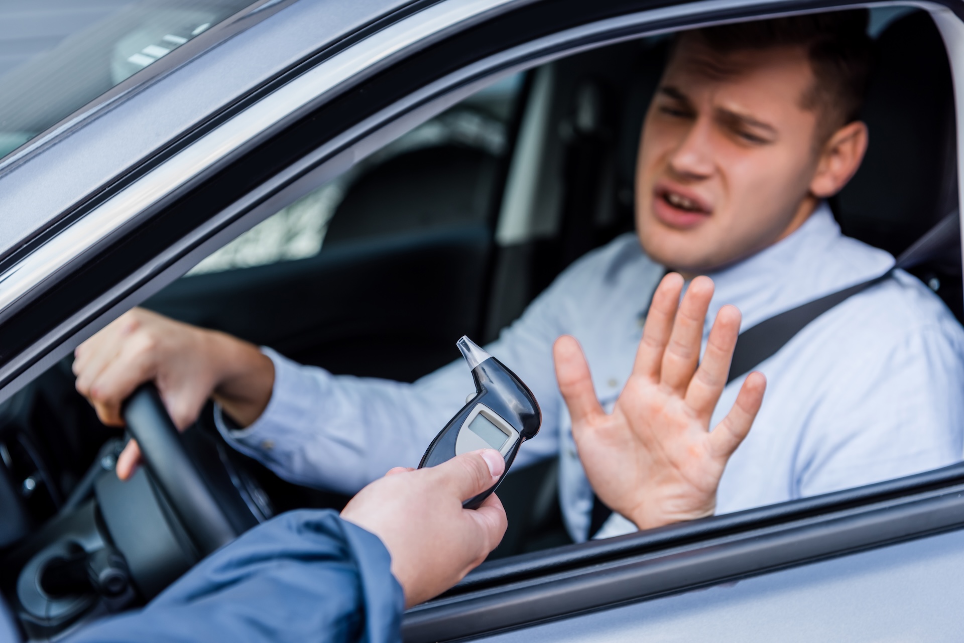 A man in a car holds up his hand, refusing to take a breathalyzer test offered by a person standing outside the vehicle.