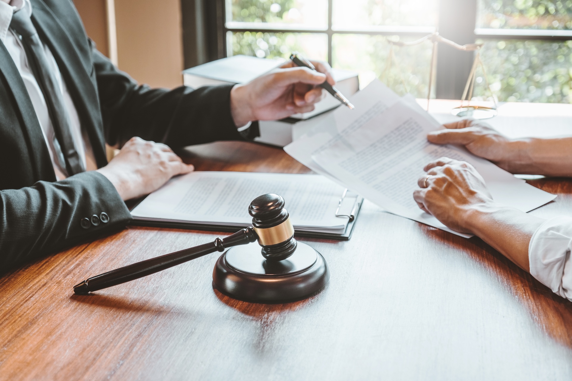 Two people sit at a wooden table, reviewing legal documents. One points with a pen, the other holds papers. A gavel and clipboard are on the table, with books and a window in the background.