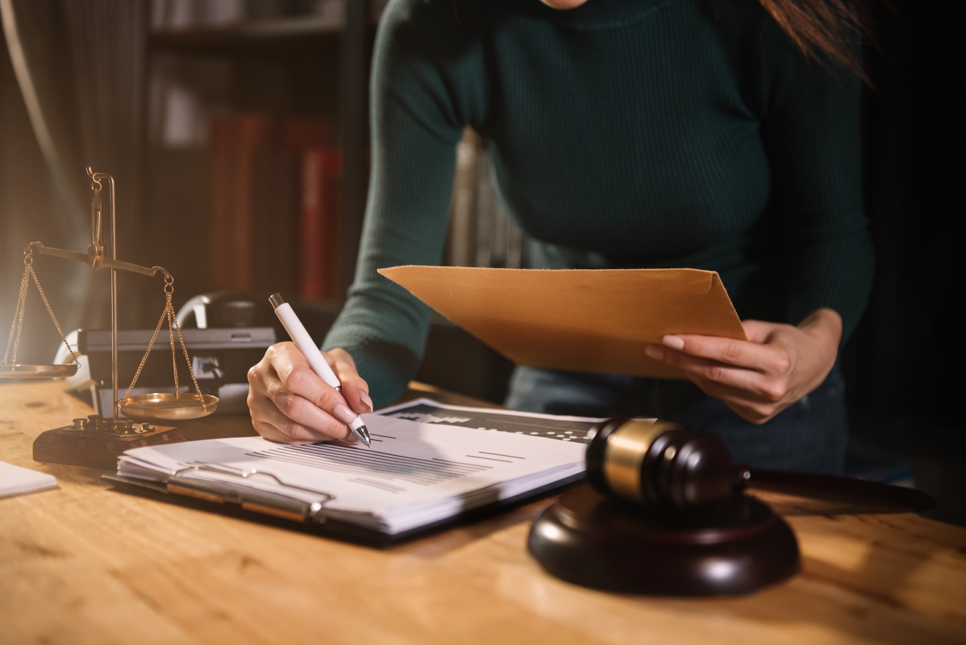 A person in a dark green sweater holds a folder and writes on documents at a desk with a gavel, scales of justice, books, and a laptop, suggesting a legal or law office setting.
