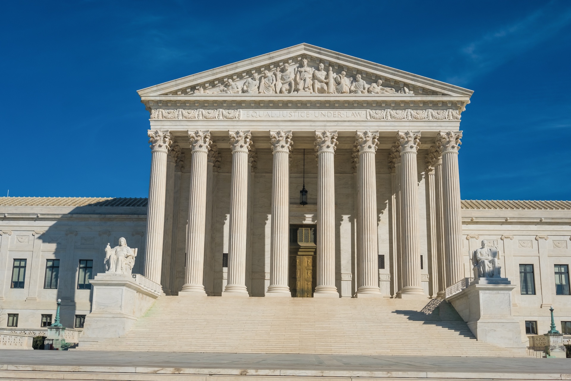 The front view of the United States Supreme Court building, featuring large columns, steps leading to the entrance, statues on either side, and a blue sky in the background.