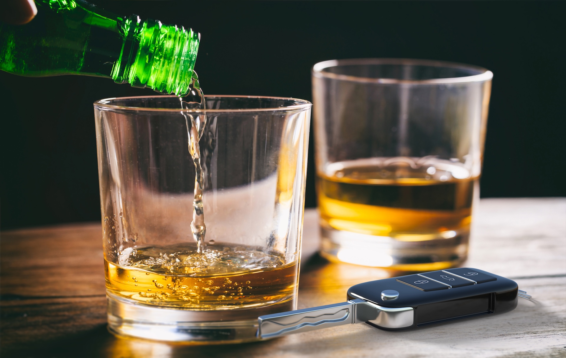 A green bottle pours alcohol into a glass on a table. Another glass with whiskey sits nearby, and a car key is placed in front, highlighting the dangers of drinking and driving.