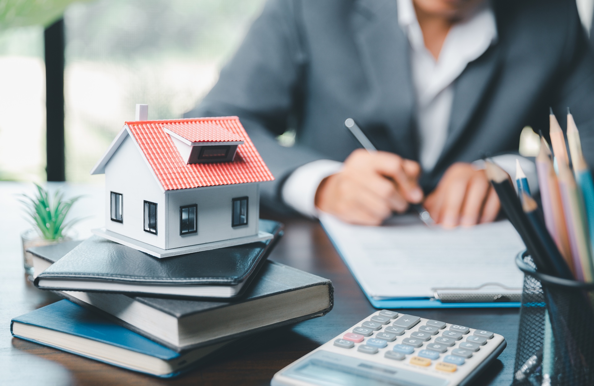 A small model house sits on top of stacked notebooks beside a calculator, as a person in a suit writes on a clipboard at a desk, suggesting a real estate or mortgage setting.