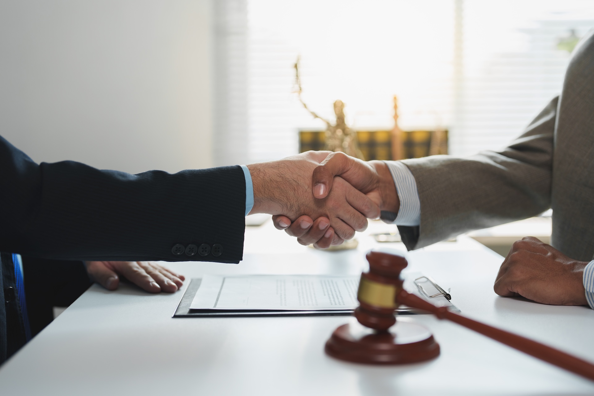 Two people in business suits shake hands across a desk with legal documents, a gavel, and a small statue of Lady Justice, suggesting a legal agreement or settlement.
