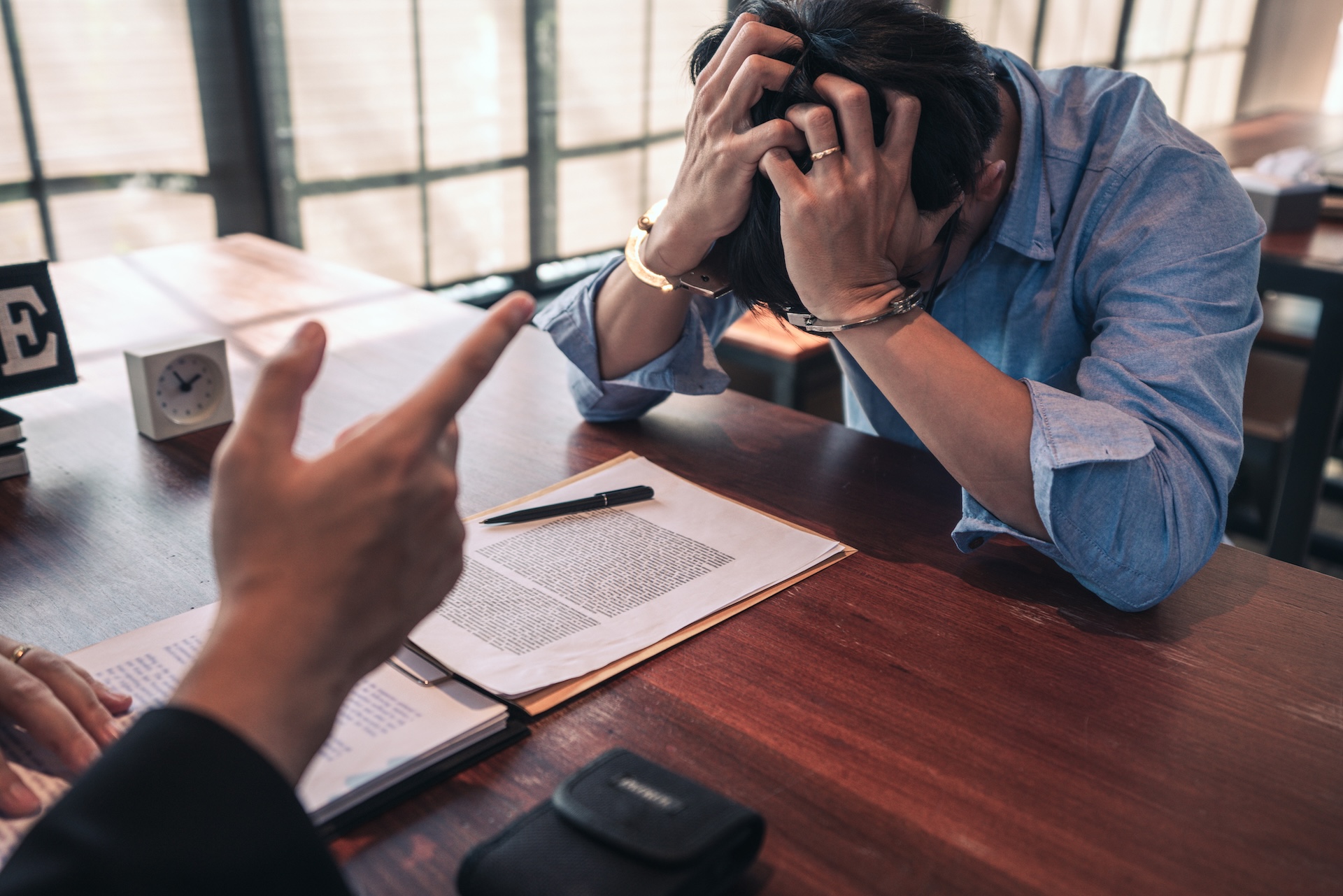 A person sits at a desk with their head in their hands, appearing stressed, while another person points at them. Papers, a pen, and a clock are on the desk in a bright room with large windows.