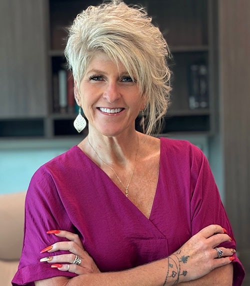 A smiling woman with short, styled blonde hair wearing a magenta top and large earrings stands with her arms crossed in an office setting. She has long red nails and several rings and tattoos on her hands.