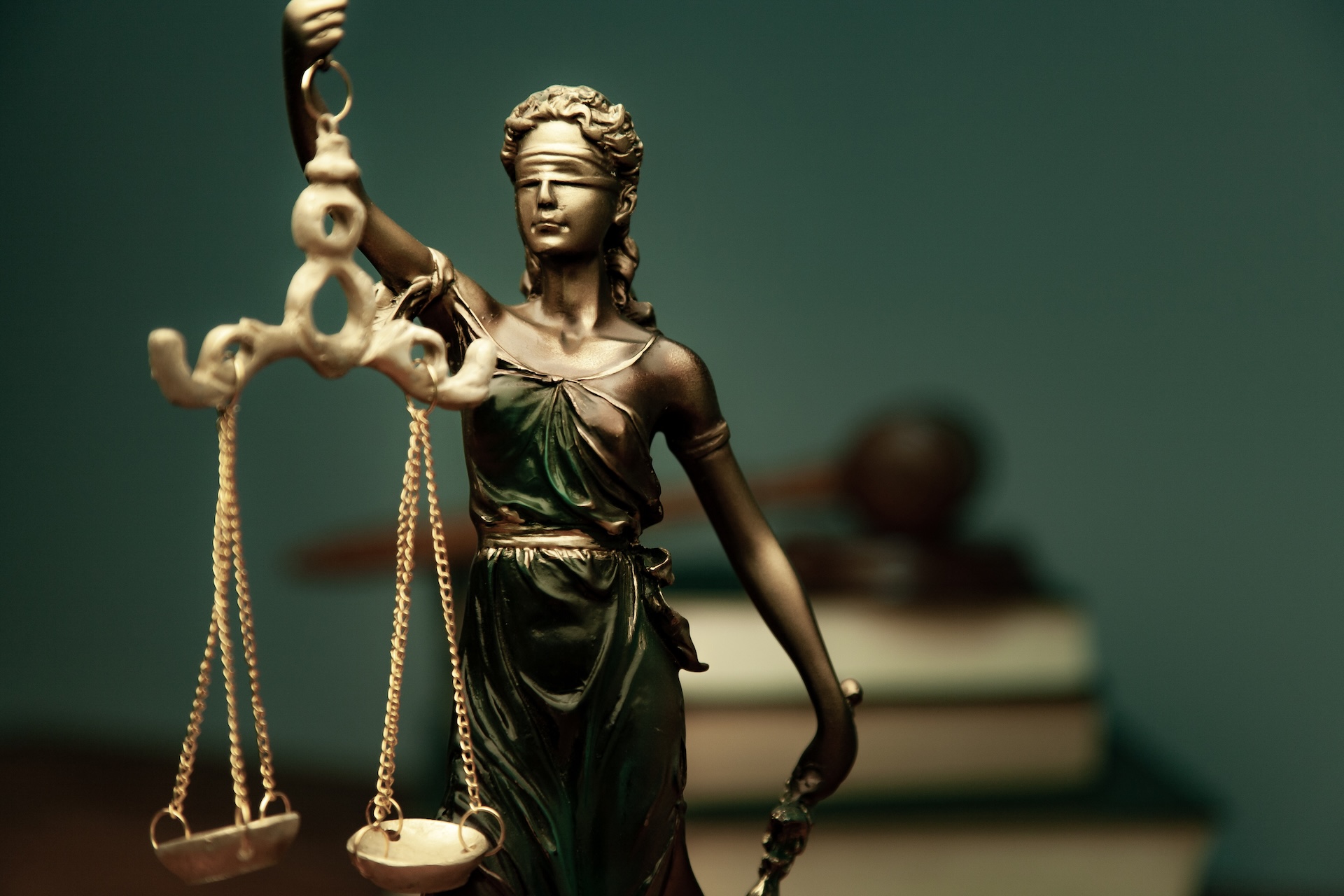 A close-up of a bronze statue of Lady Justice holding scales, wearing a blindfold. In the blurred background, a judge’s gavel rests on stacked books, symbolizing law and justice.