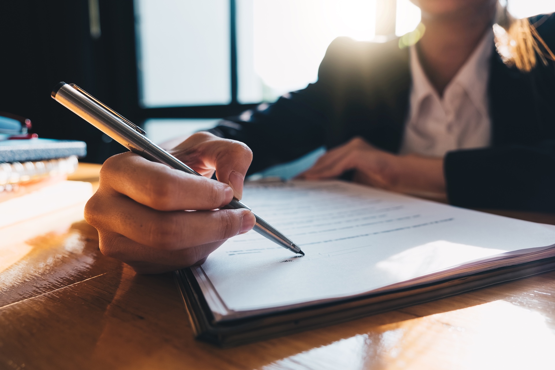 A person in business attire sits at a desk, holding a pen and signing a document, with sunlight streaming through a window in the background.