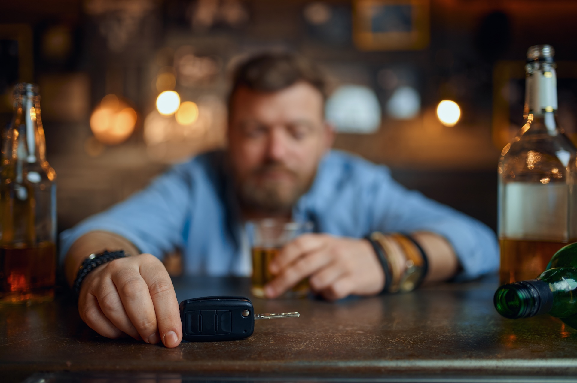 A man with a beard sits at a bar holding a glass of alcohol, with several bottles around him. His other hand rests on a set of car keys on the counter, suggesting the dangers of drinking and driving.