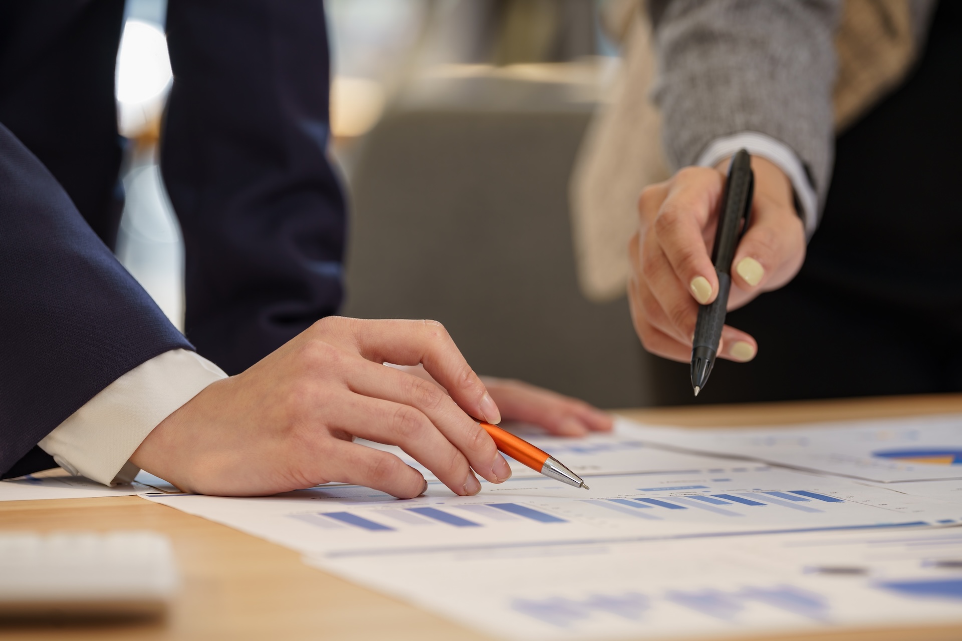 Two people reviewing printed charts and graphs on a table, one holding an orange pen and the other pointing with a black pen, suggesting discussion or analysis of financial or business data.