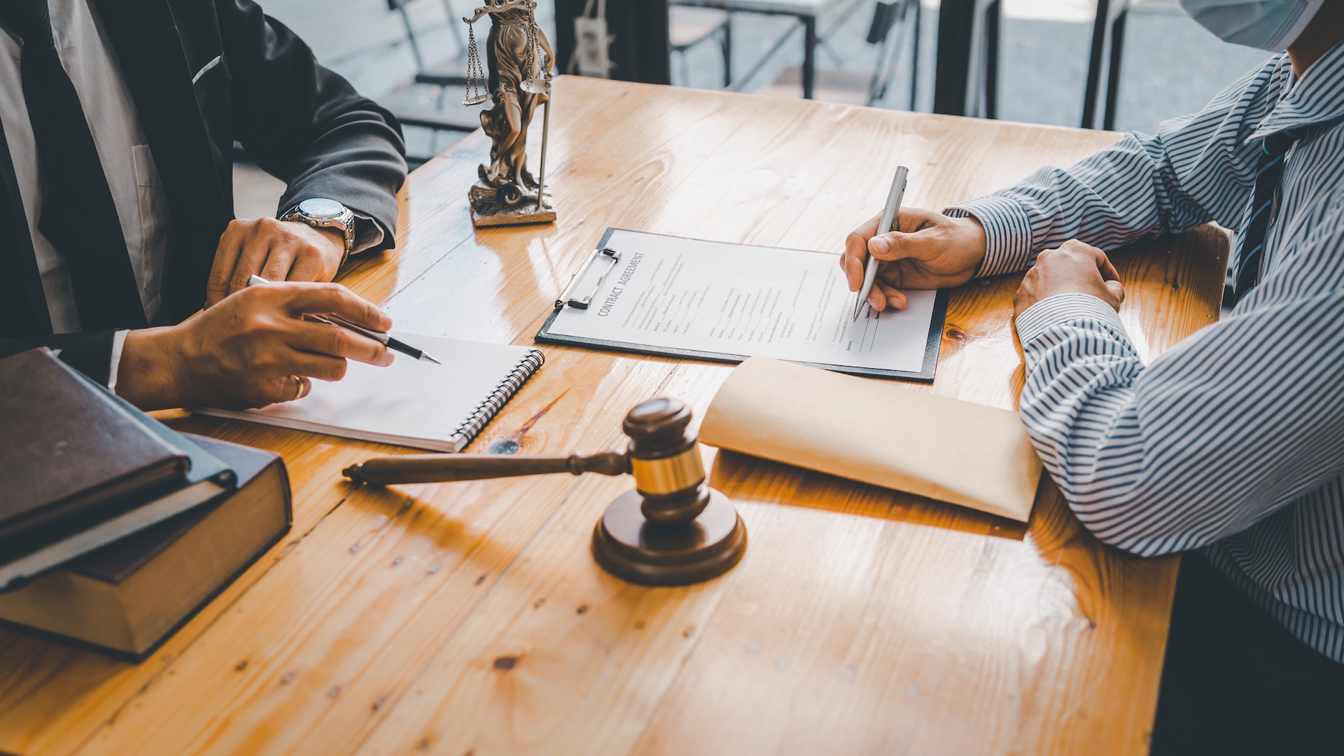 Two people sit at a wooden table with legal documents, a gavel, law books, and a small Lady Justice statue, suggesting a legal consultation or meeting. One person points while the other writes on a clipboard.