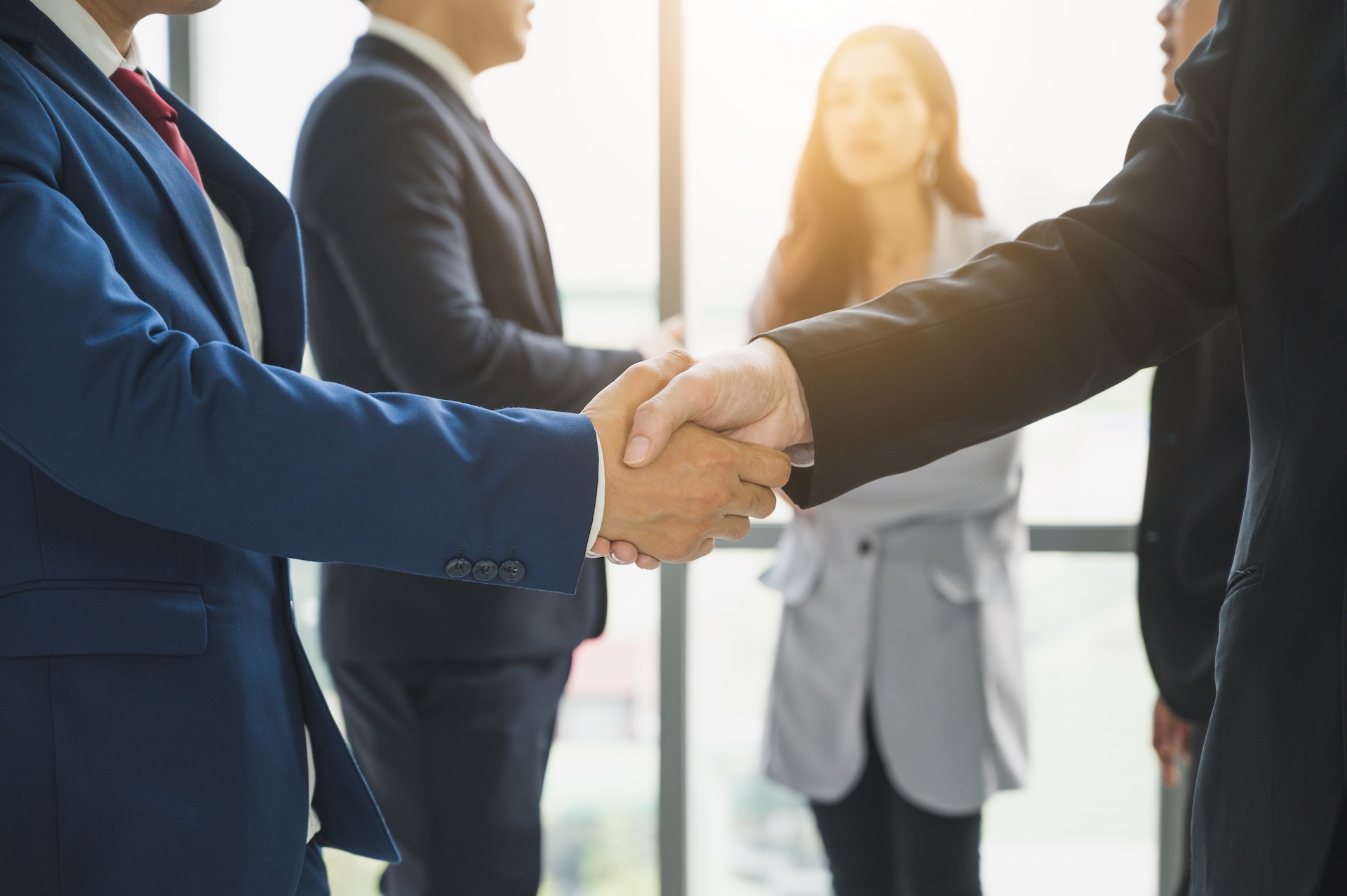 Two people in business suits shake hands in the foreground, representing a successful partnership, while three other professionals stand in the background of a bright office—an ideal setting for discussing the Houston Estate Planning and Probate Guide.