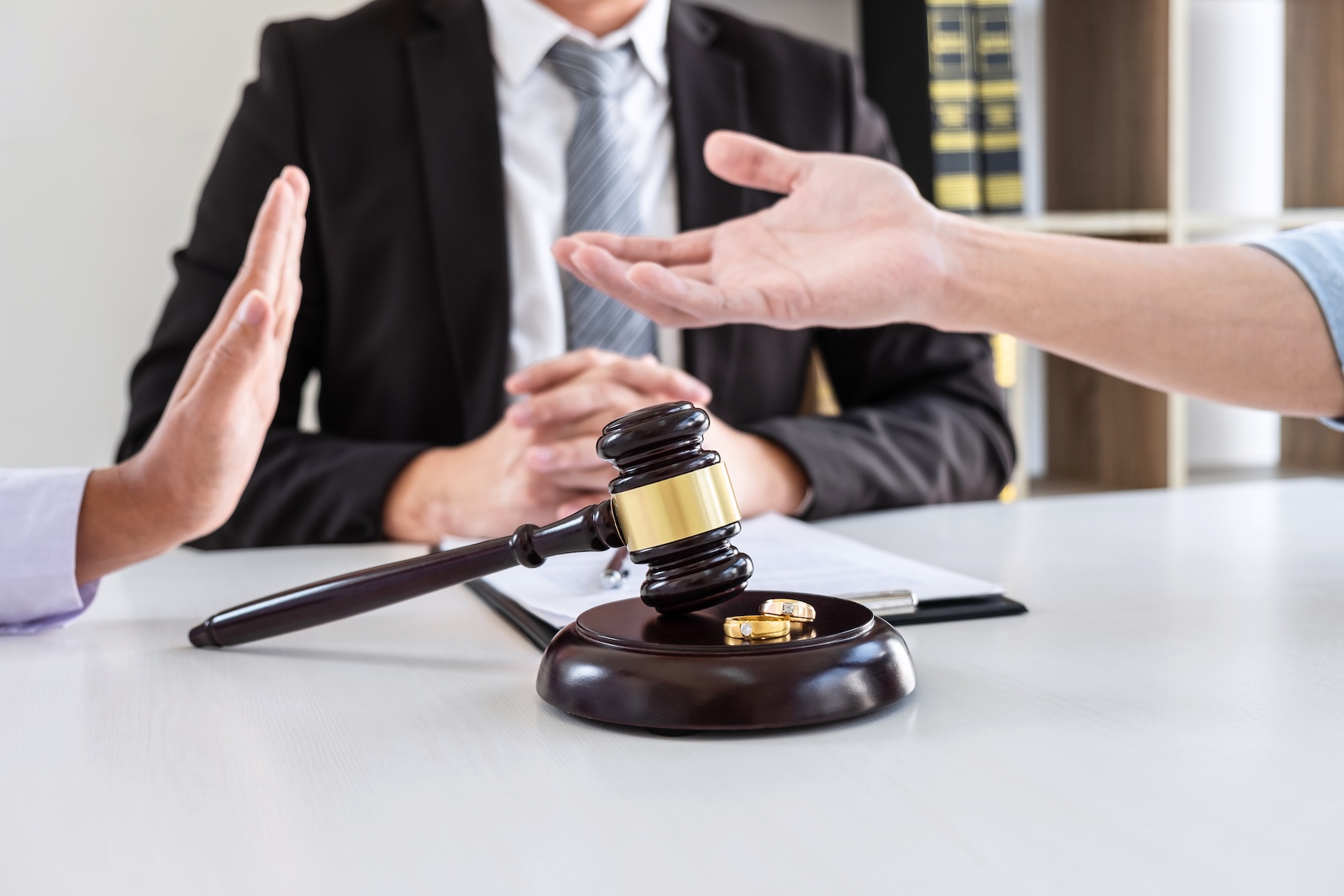 Two people gesture emotionally across a table with a judge’s gavel and wedding rings between them, while a person in a suit sits in the background, suggesting a legal or divorce proceeding.