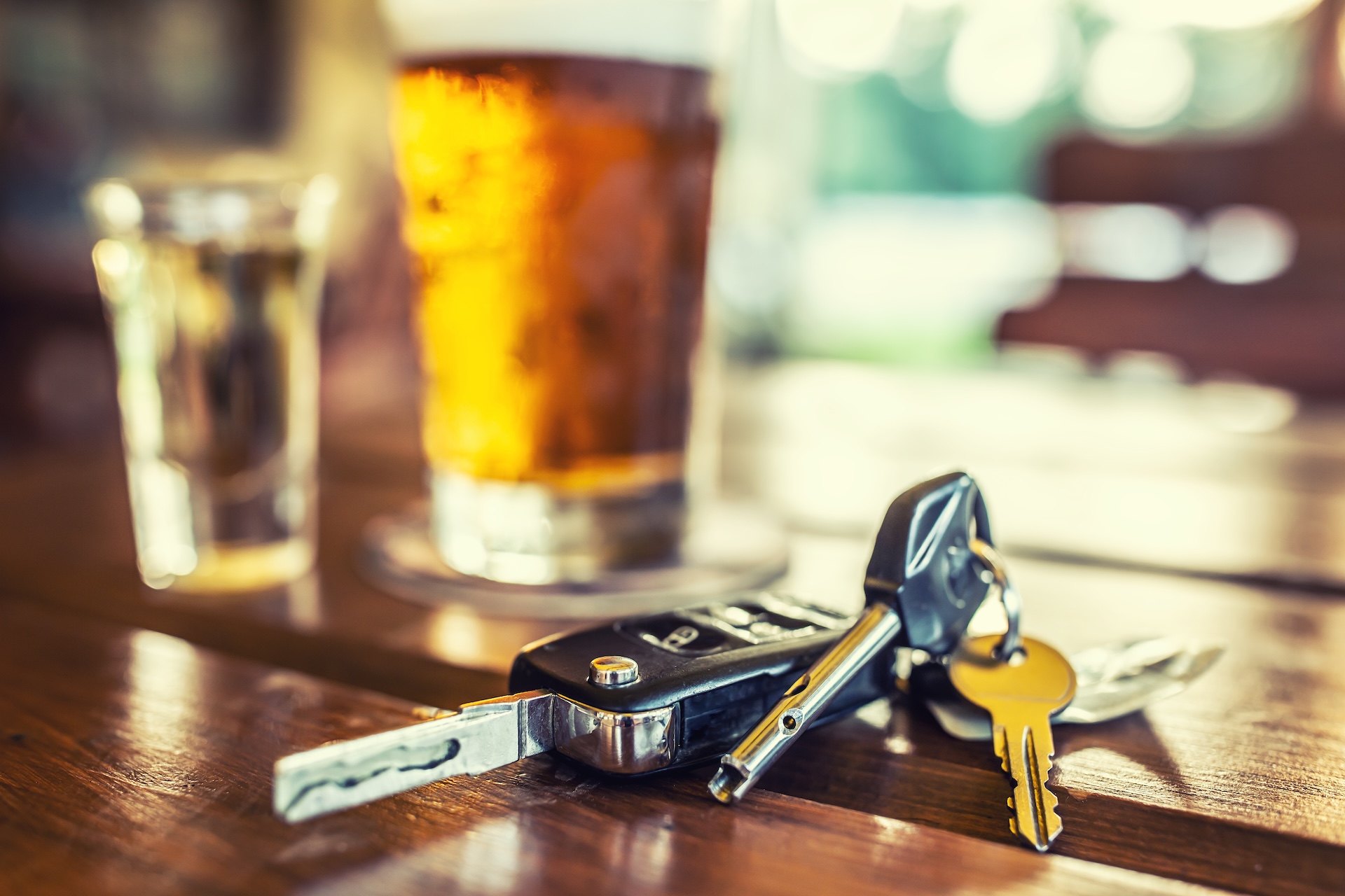 A set of car keys on a wooden table with a glass of beer and a shot glass in the background, highlighting the risks of drinking and driving. Arrested for DWI in Montgomery County? Here’s what you need to know.