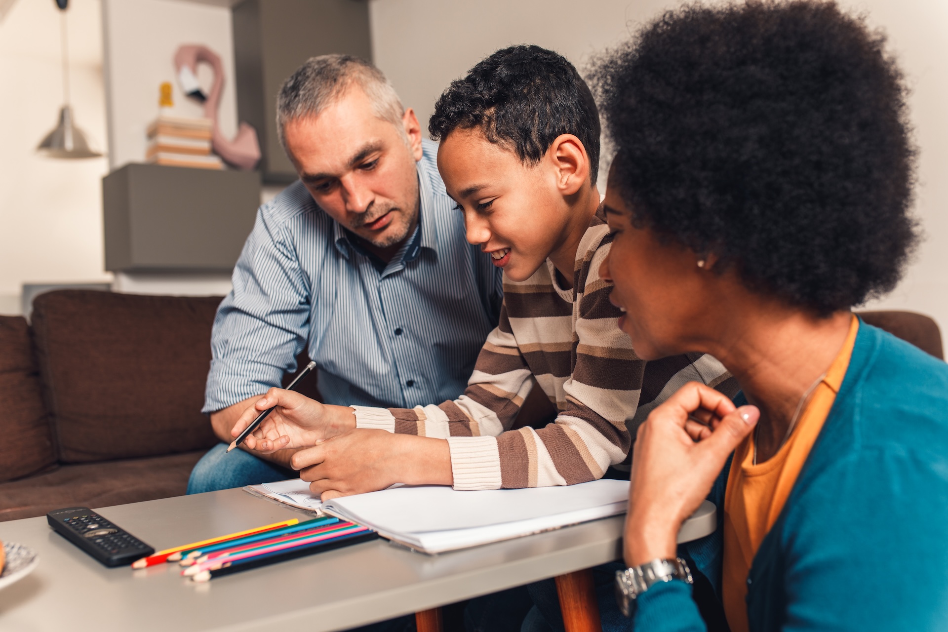A boy sits at a table with an open notebook, smiling and holding a pencil, while an adult man and woman—perhaps following an Extended Standard Possession Order Texas—help with homework. Colored pencils and a remote are on the table.