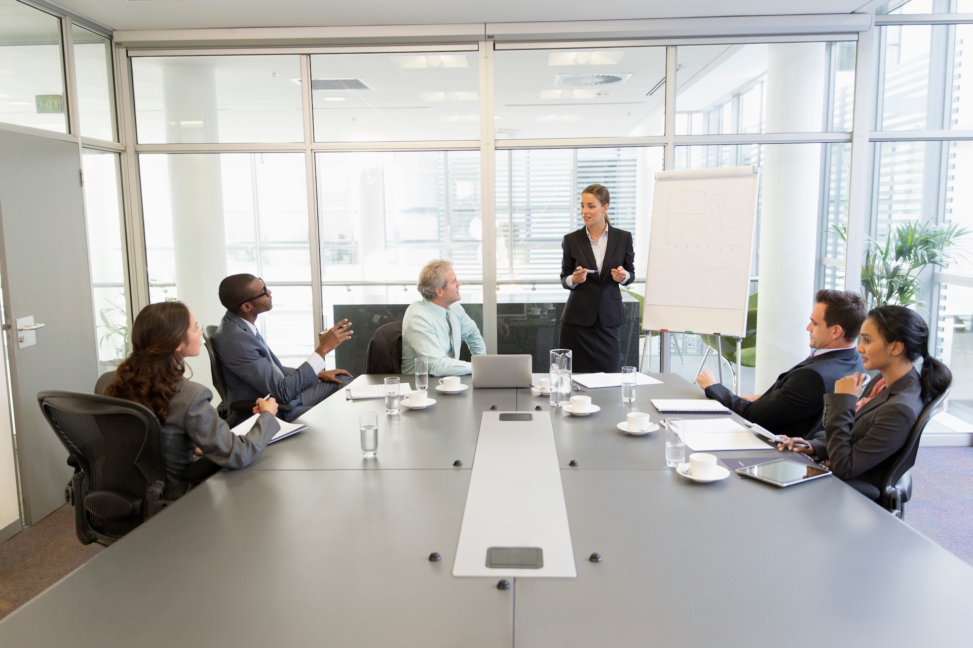 Five people sit at a conference table while a woman stands by a flip chart, presenting to the group about the Corporate Transparency Act BOI. Laptops, papers, coffee cups, and glasses of water are on the table in a modern Texas office setting.