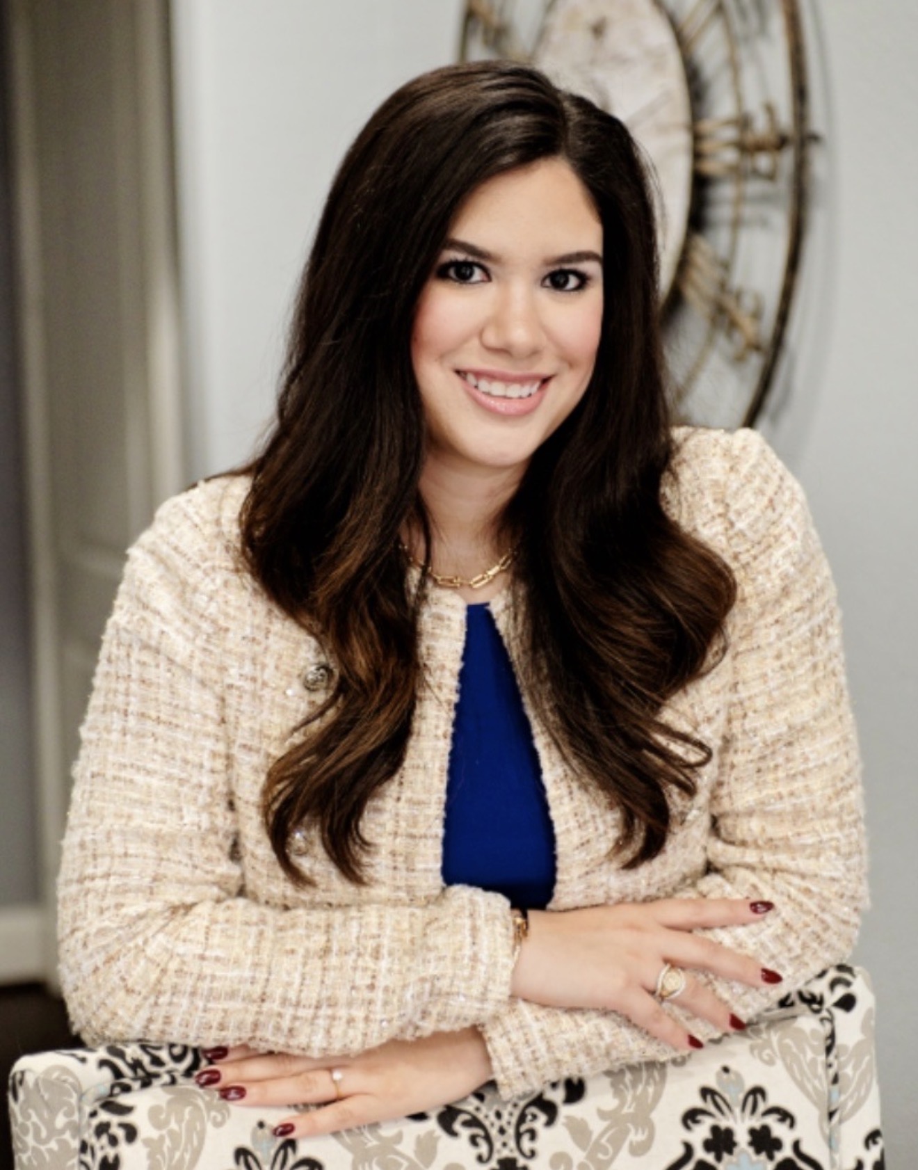A woman with long dark hair, wearing a cream textured jacket over a blue top, sits and smiles with her arms crossed on a patterned chair. A decorative wall clock is visible in the background.