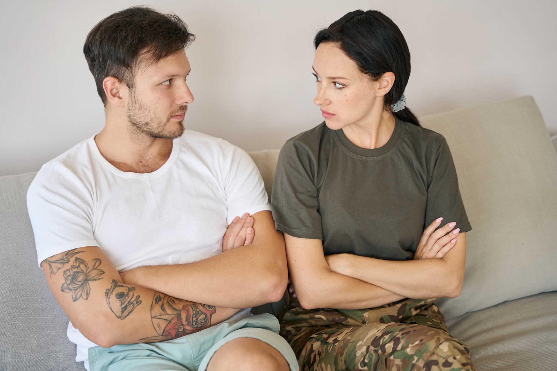 A man and woman sit on a couch with arms crossed, facing each other with serious expressions—perhaps discussing a military divorce in Texas. The man wears a white shirt and shorts; the woman wears a green shirt and camouflage pants.