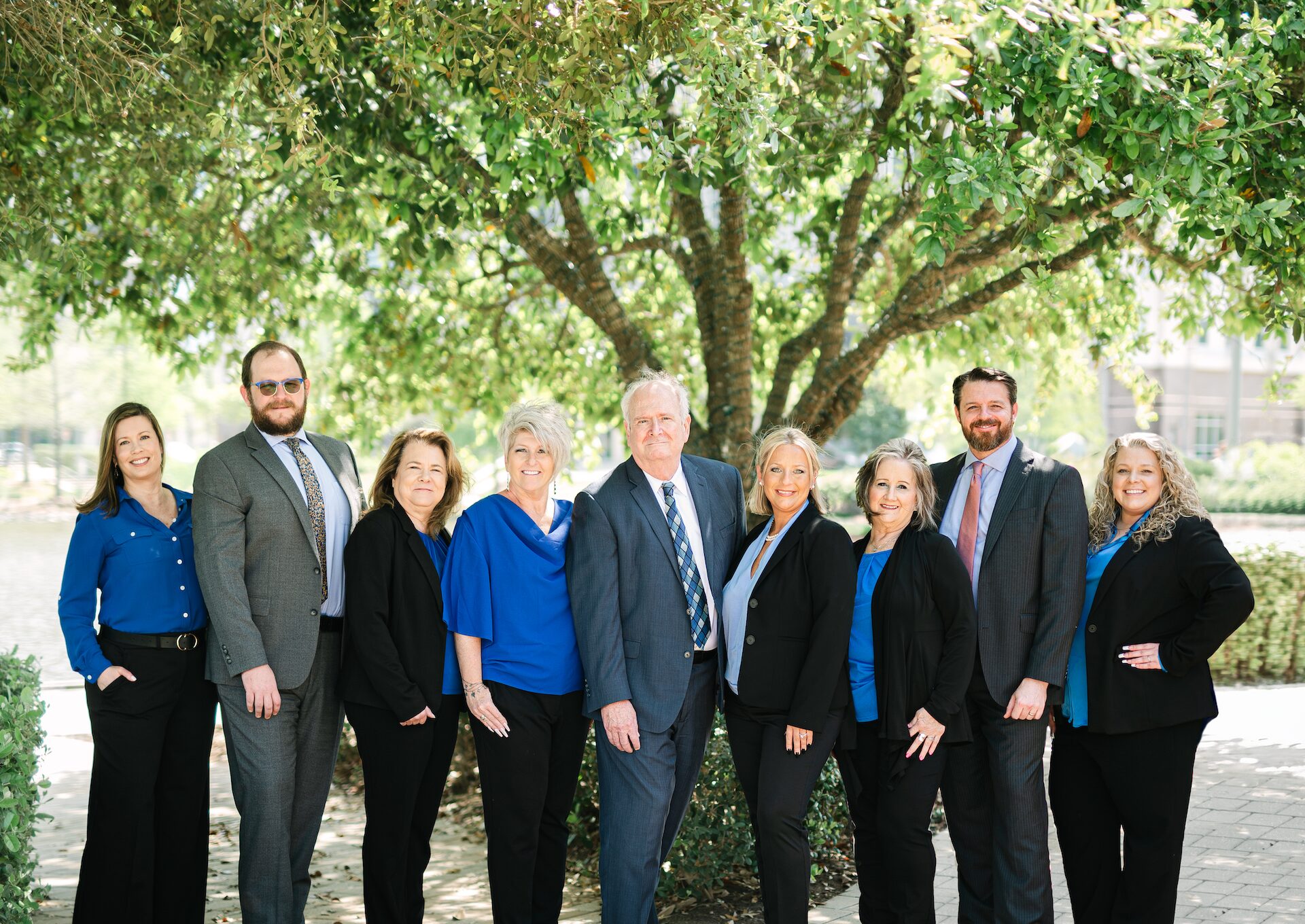 Nine professionally dressed people stand together outside beneath a large tree, smiling at the camera. The group includes both men and women, all wearing blue and black attire, with greenery and sunlight in the background. Nine professionally dressed people stand together outside beneath a large tree, smiling at the camera. The group includes both men and women, all wearing blue and black attire, with greenery and sunlight in the background.