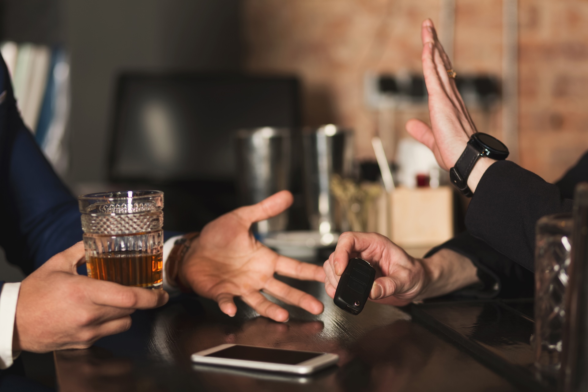 A person holding a glass of alcohol offers car keys to another, who refuses with a raised hand gesture—a reminder to stay safe and follow Texas DWI laws 2026. A smartphone sits on the bar nearby.
