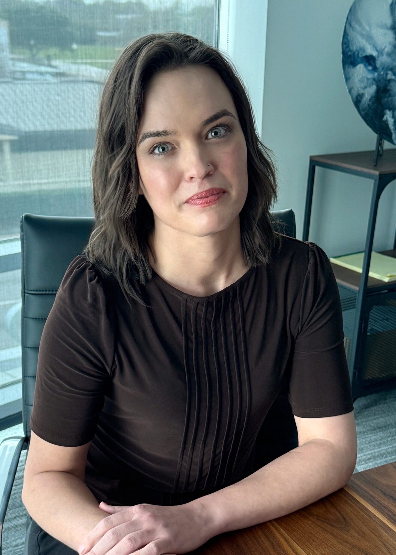 A woman with shoulder-length brown hair and green eyes sits at a wooden desk in an office, wearing a short-sleeved black blouse. There is a window and a globe on a stand in the background.