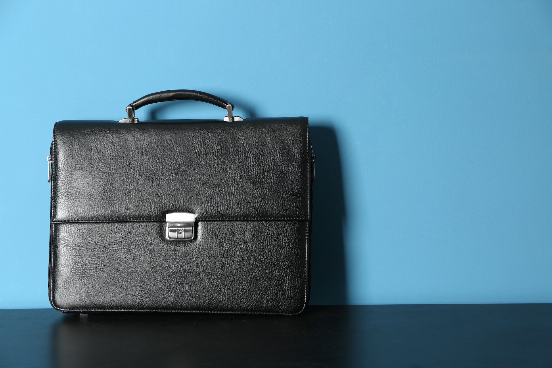 A black leather briefcase with a handle and a silver clasp sits on a black surface against a plain blue wall, evoking the professional decisions faced when considering LLC vs corporation Texas.