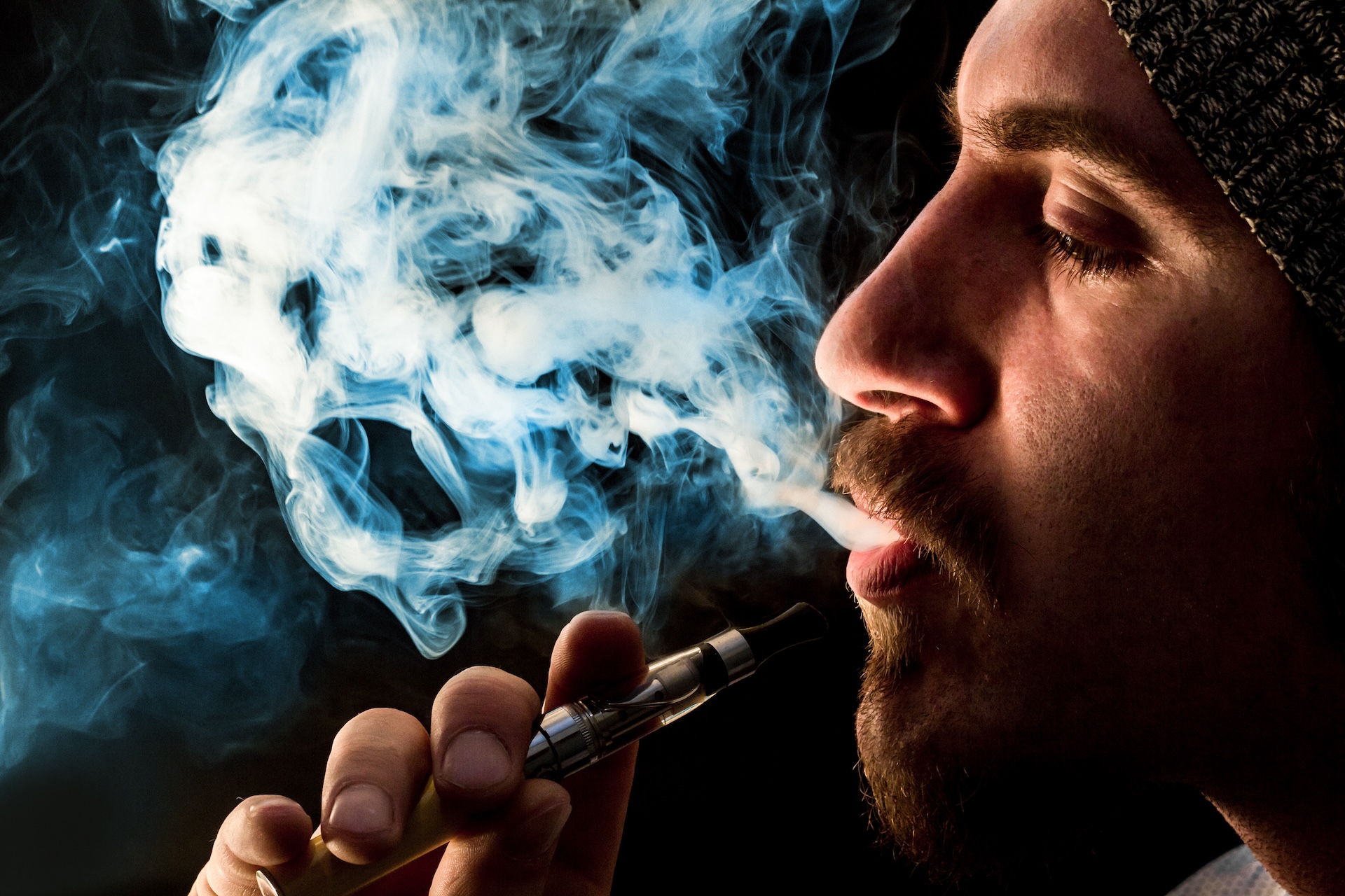 A person wearing a knit hat exhales a thick cloud of vapor while holding a vape device, with swirling smoke visible against a dark background—a scene that may soon disappear with the Texas vape ban 2025 approaching.