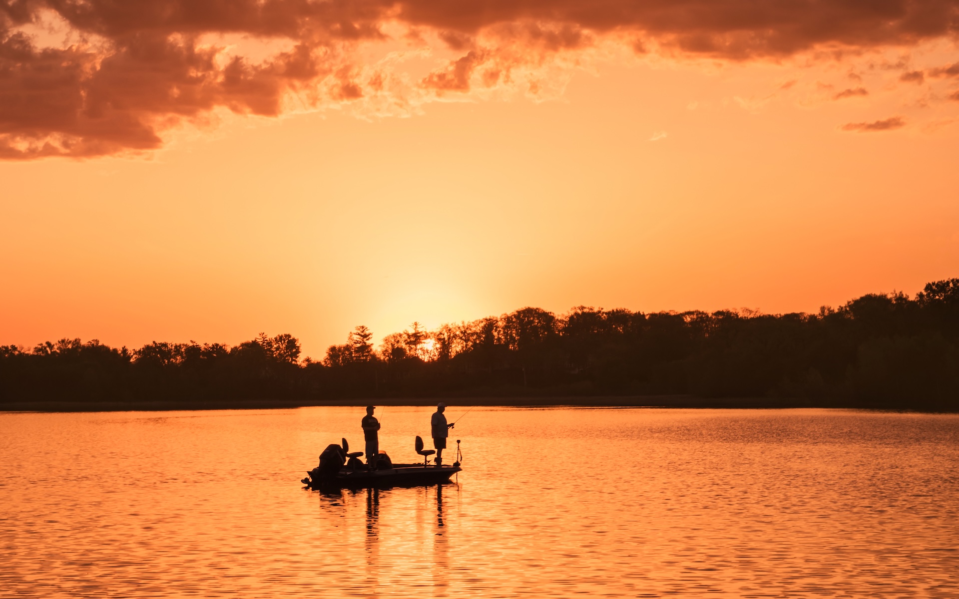 Two people are fishing from a small boat on a calm lake at sunset. The sky and water glow with warm orange and yellow hues, and trees are silhouetted in the background.