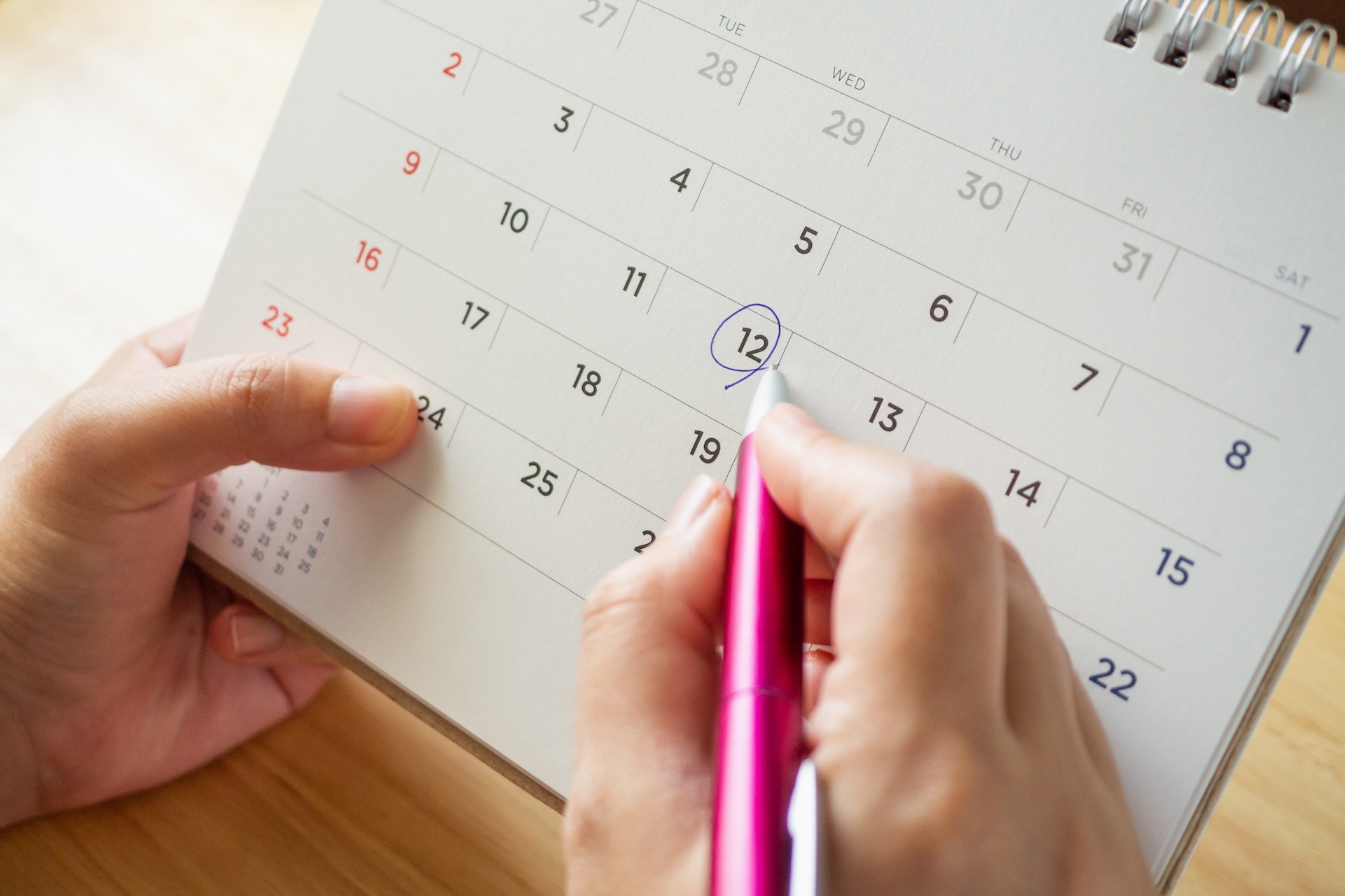 A hand holding a pen marking a date on a paper desk calendar.