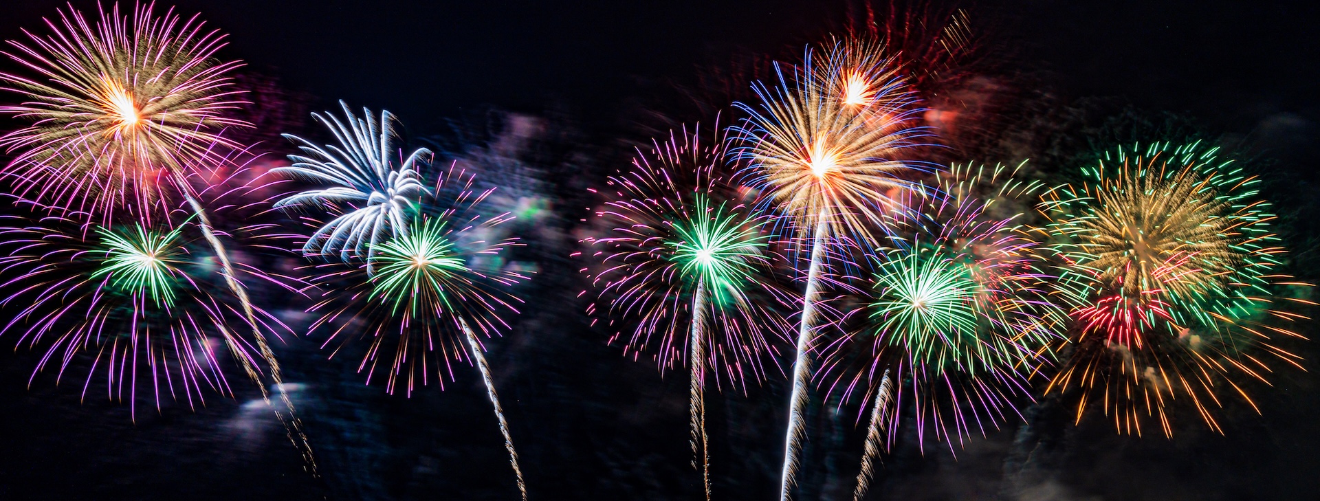 Colorful fireworks burst in the night sky, displaying vibrant shades of pink, purple, green, yellow, blue, and orange with long trails and radiant sparks.