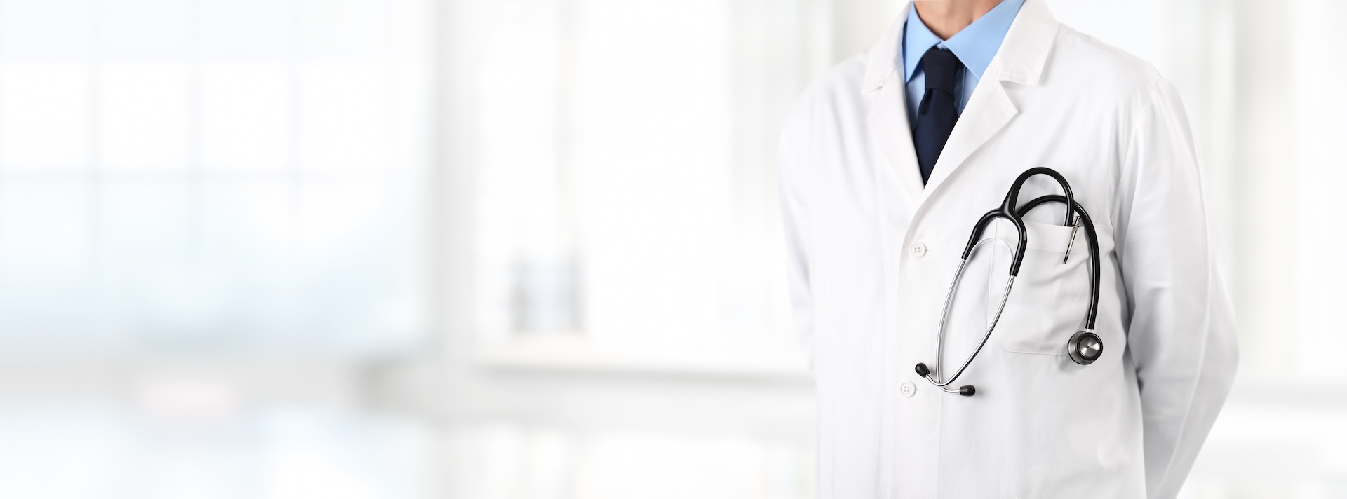 A doctor wearing a white lab coat and a stethoscope around their neck, standing in a bright, blurred medical office. The person’s face is not visible.