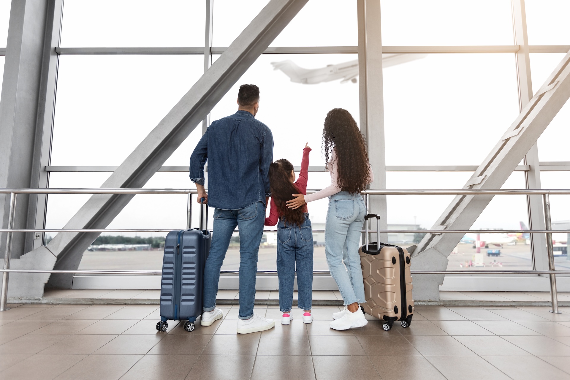 A family of three stands in an airport terminal with suitcases, looking out a large window. The child points at an airplane taking off outside. All are seen from behind.