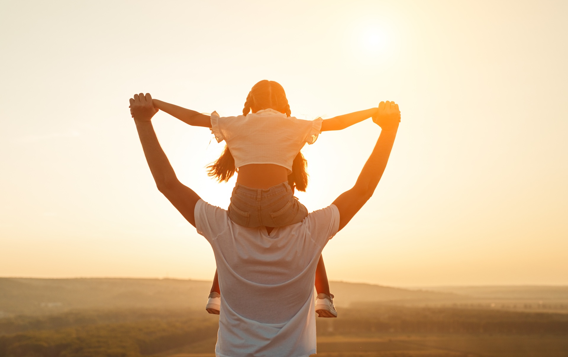 A father lifting his young daughter onto his shoulders against a warm sunset sky.