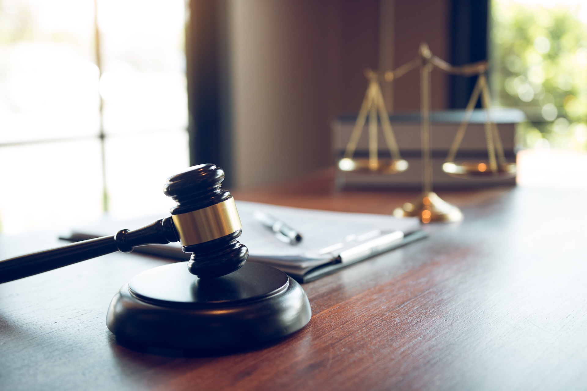 A wooden judge's gavel rests on a table beside a clipboard with papers and a pen, with a blurred scale of justice in the background near a window.