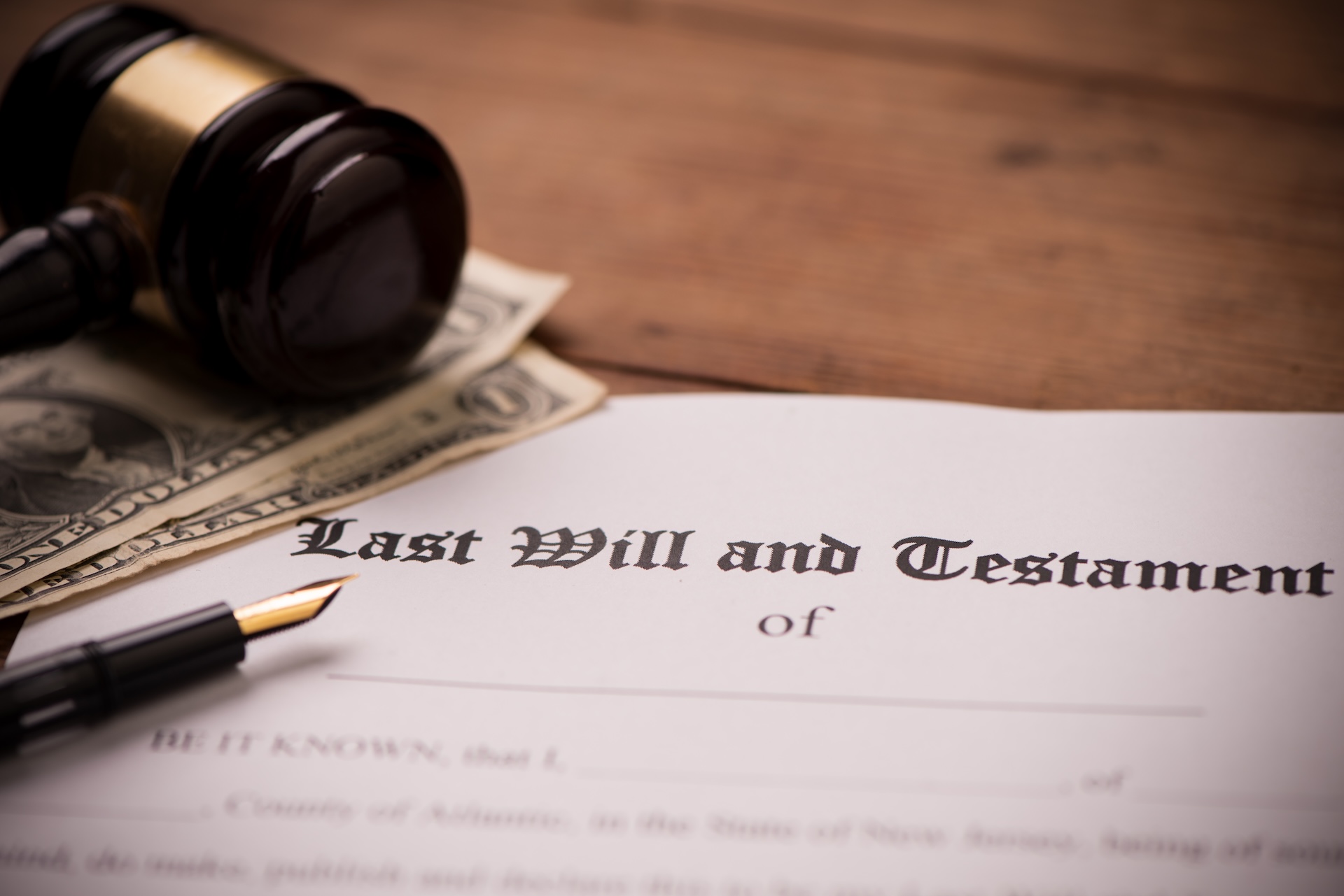 A last will and testament form sits on a wooden table next to a fountain pen, a judge’s gavel, and two one-dollar bills.