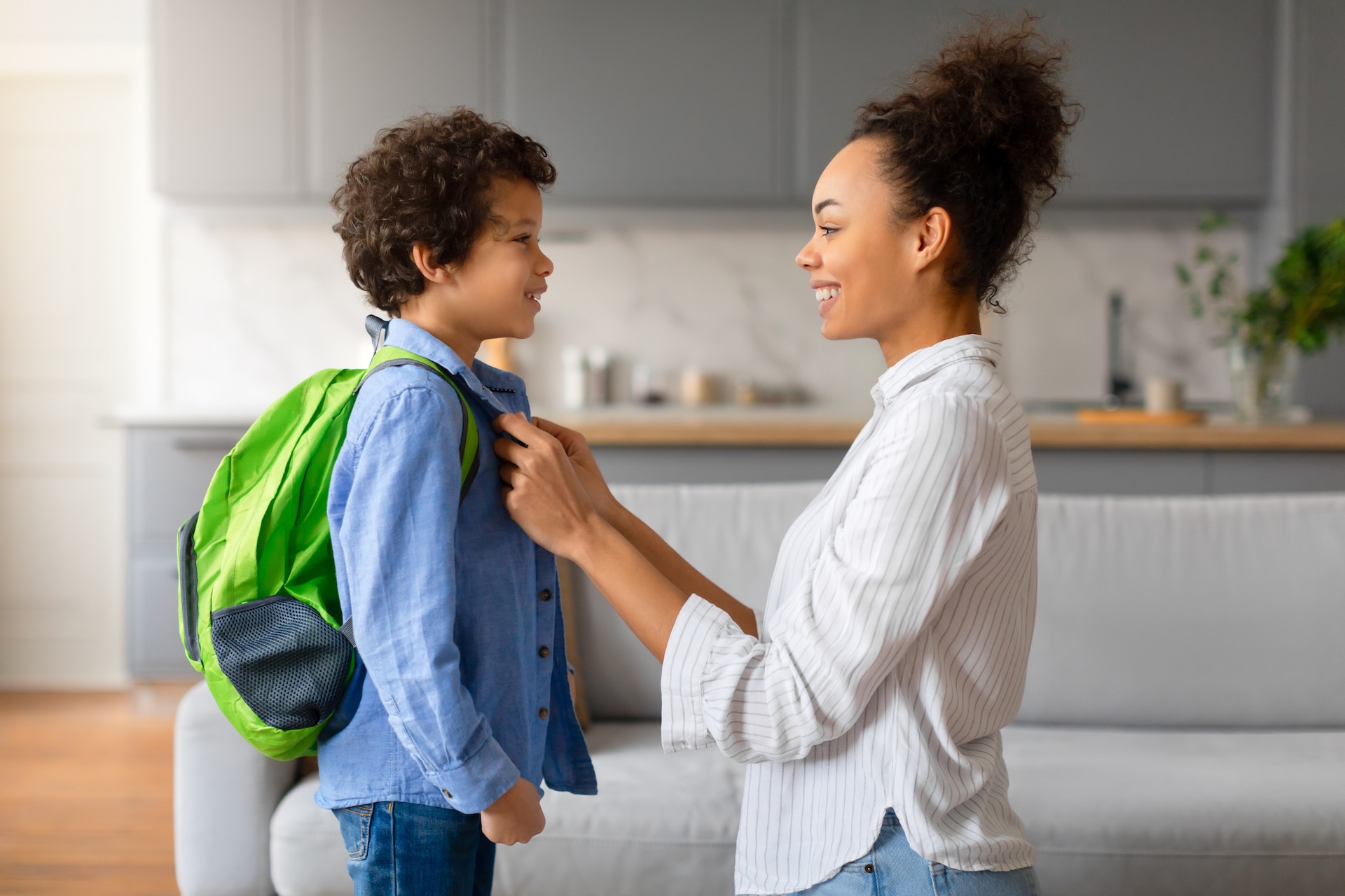 A woman smiles while adjusting the collar of a young boy wearing a green backpack. They are standing in a bright living room with a grey sofa and a kitchen in the background.