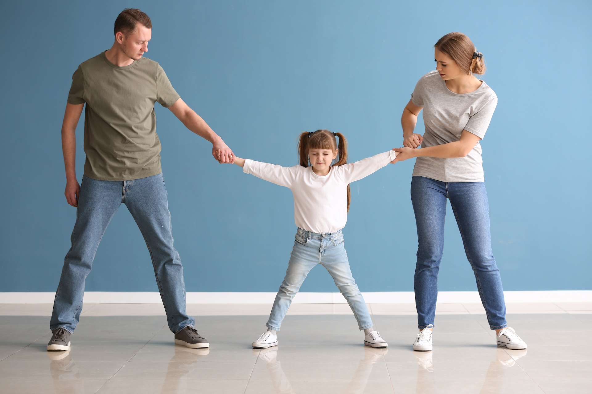 A young girl stands between a man and a woman, each holding one of her hands and pulling her in opposite directions. They all look serious. The scene takes place indoors against a blue wall.